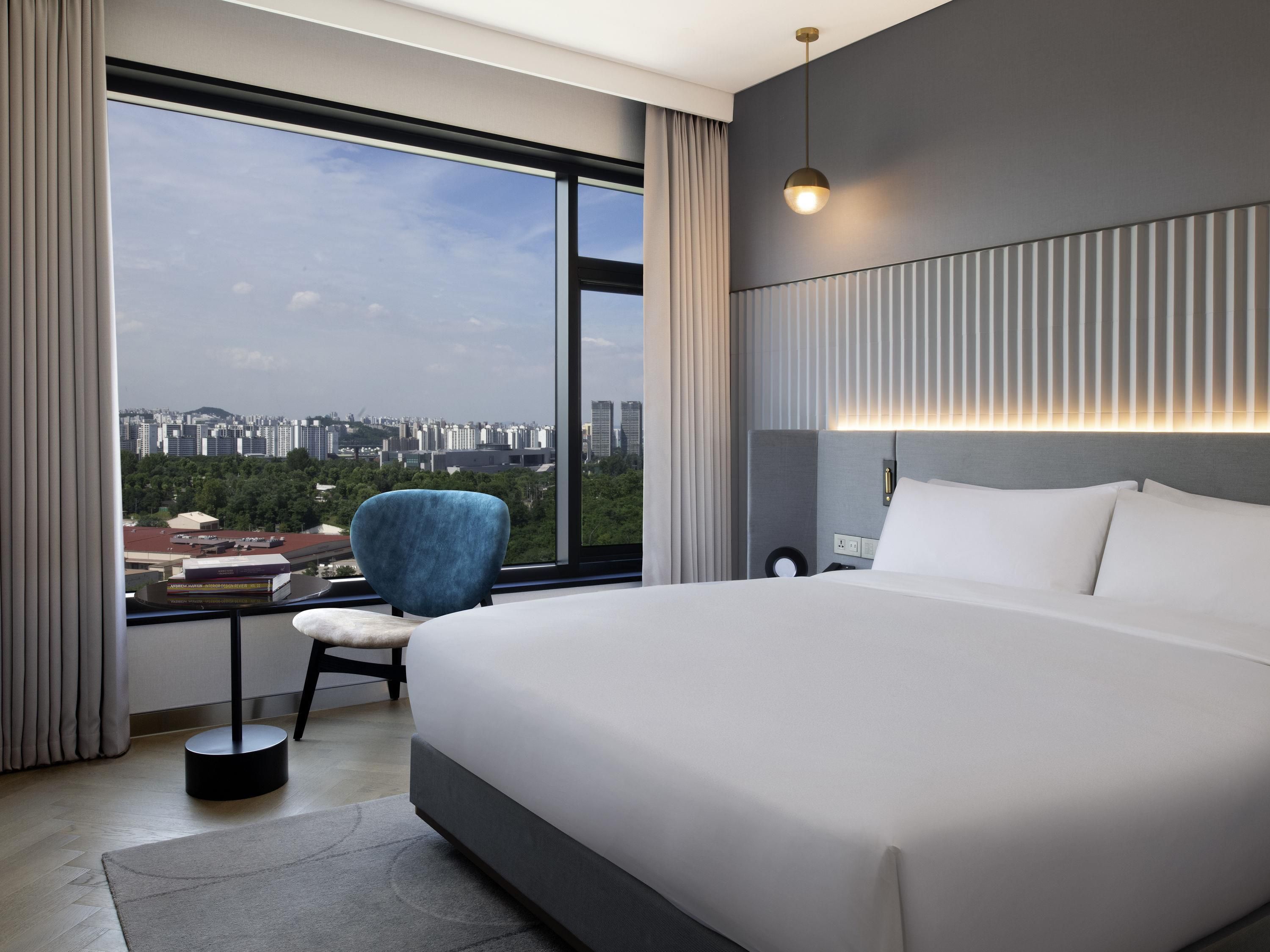 A modern hotel room with a large bed, a chair, a bedside table, and a view of a cityscape through large windows.