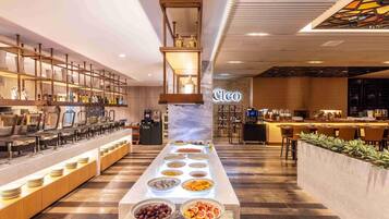 A modern hotel lobby with a long buffet table, a coffee station, and a seating area with a fireplace.