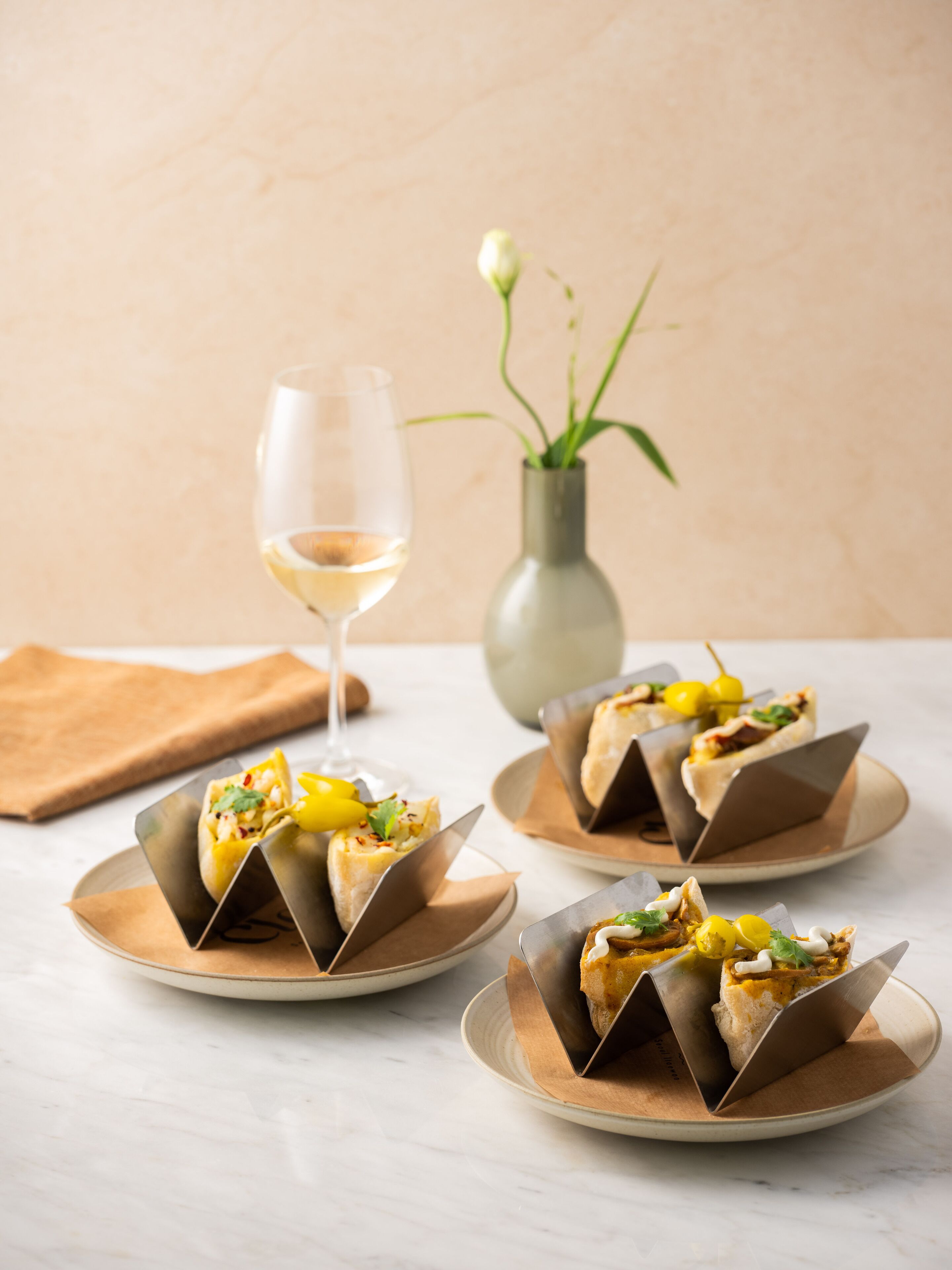Three plates of food with a glass of white wine and a vase with flowers in the background.