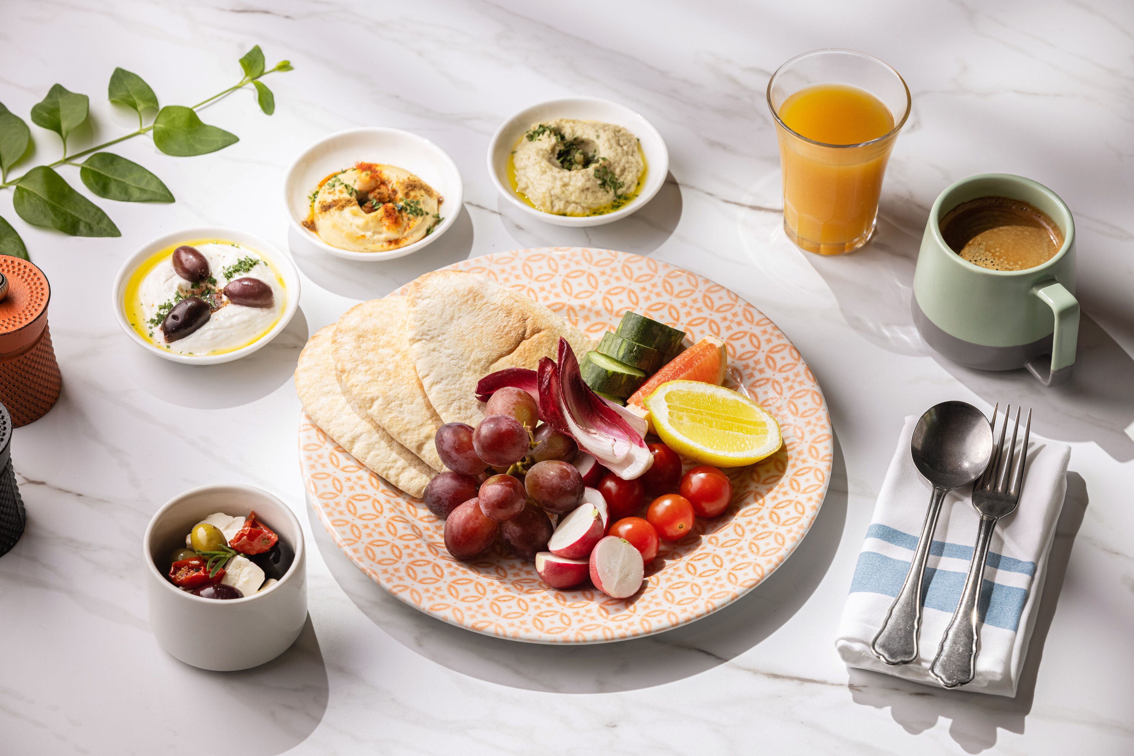 A table with a plate of food, including pita bread, grapes, tomatoes, and olives, along with bowls of hummus and a glass of orange juice.