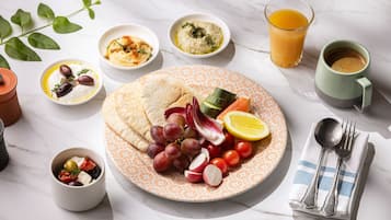 A table with a plate of food, including pita bread, grapes, tomatoes, and olives, along with bowls of hummus and a glass of orange juice.