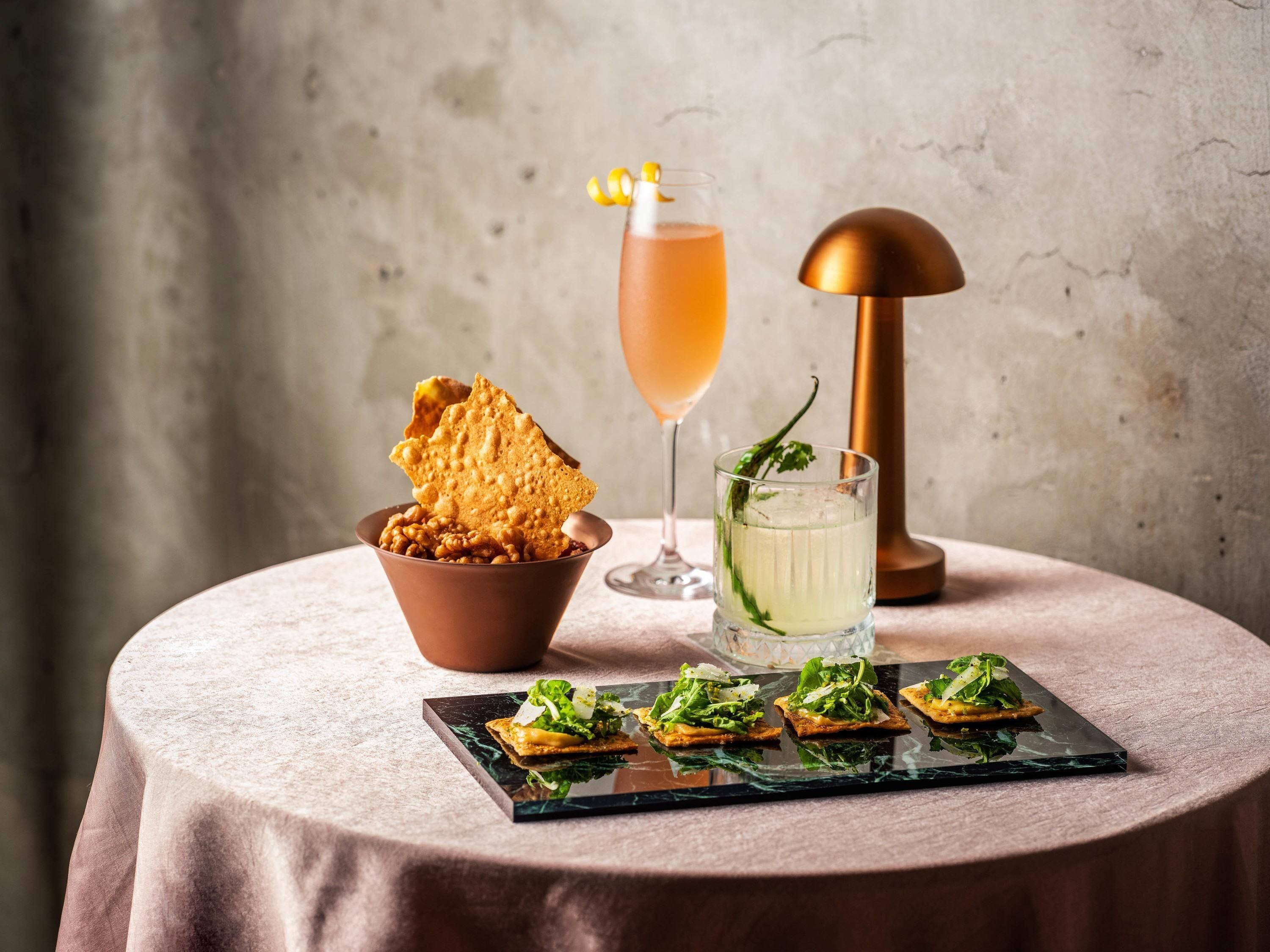 A table with a bowl of nuts, a glass of drink, a lamp, and a plate of appetizers.