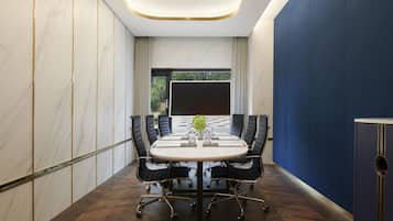 A conference room with a round table, black chairs, a large screen, and a view of greenery outside.