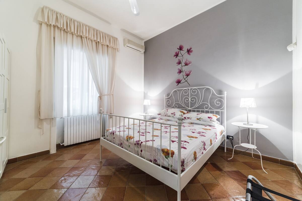 A bedroom with a bed, a radiator, a window with curtains, a wall-mounted air conditioner, and a small table with a lamp.