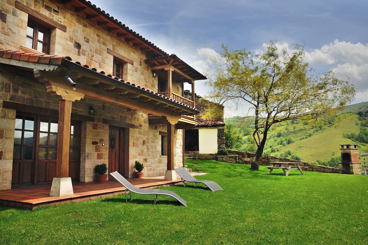 Una casa de piedra con un balcón, un césped con dos sillones de jardín y una mesa de picnic.