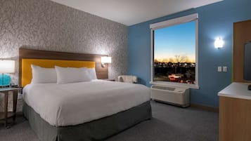 A hotel room with a large bed, a window with a city view, a desk with a lamp, and a heater.
