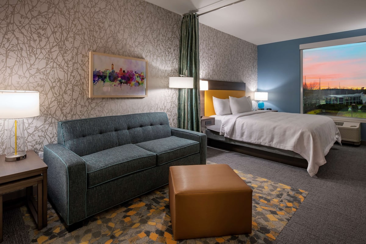 Ein Hotelzimmer mit einer Couch, einem Bett, einem Nachttisch, einer Lampe, einem Gemälde und Blick auf den Sonnenuntergang.