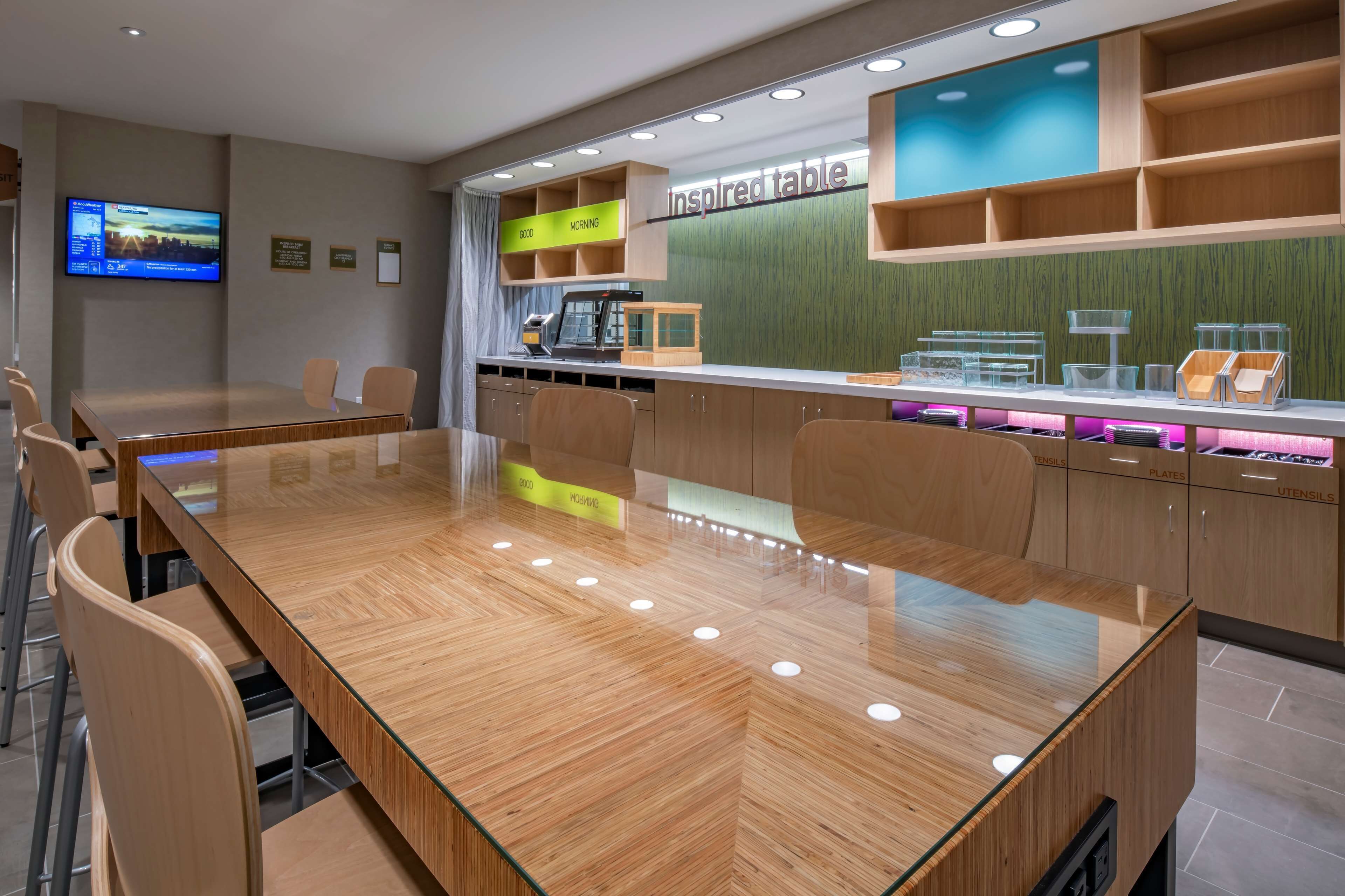 A conference room with a glass-top table, wooden chairs, a television on the wall, and a counter with labeled compartments for utensils and plates.