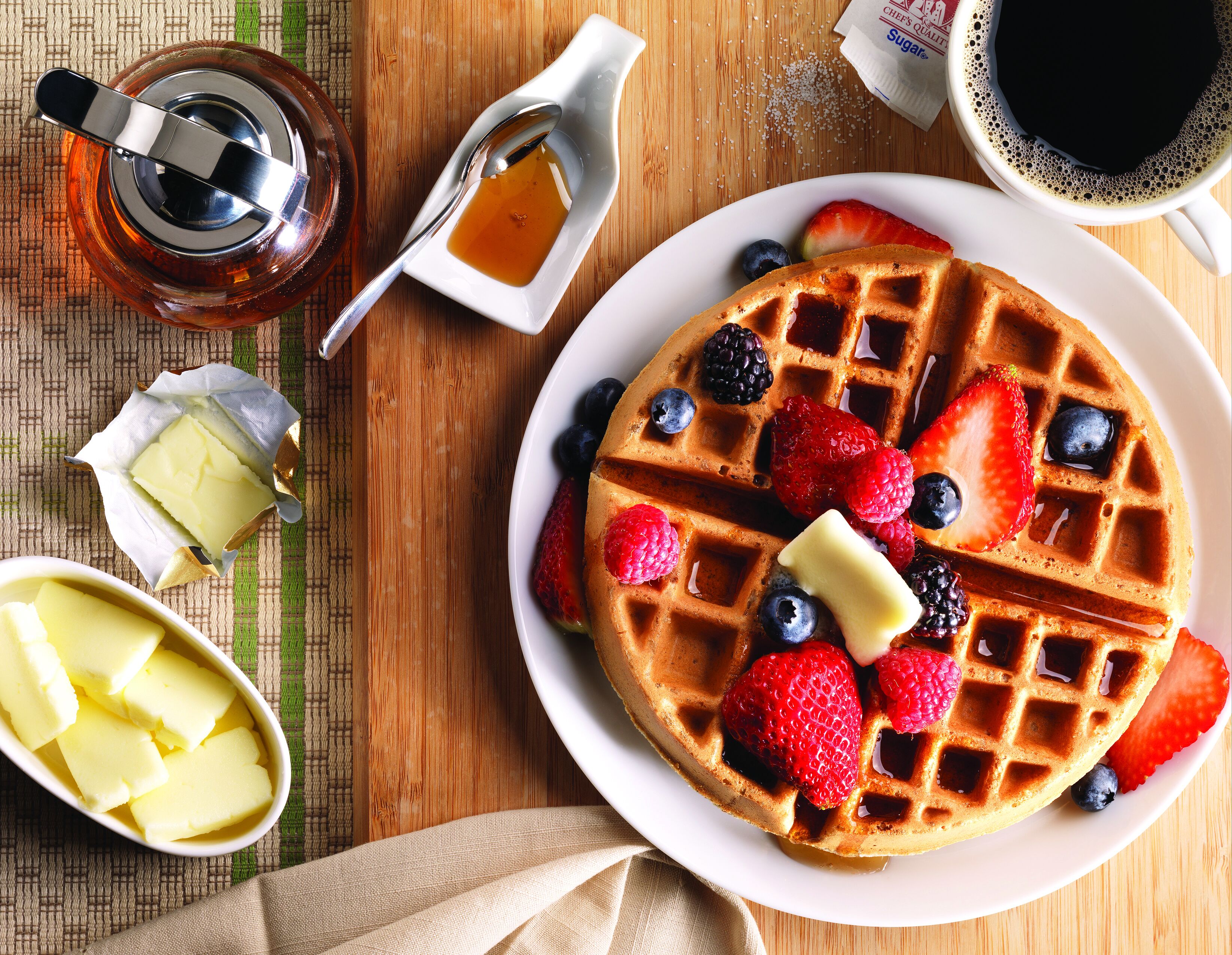 Une gaufre garnie de fraises, de myrtilles et de mûres, servie avec du beurre et du sirop.