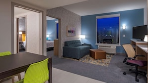 A hotel room with a dining area, a sofa, a TV, and a window with a city view. - Home2 Suites by Hilton Wayne, NJ (Wayne)
