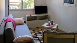 Una sala de estar con sofá, televisión, una mesita con bebidas, una lámpara y una ventana con vista a un edificio.