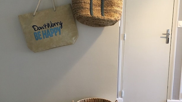 Una habitación con un banco de madera, una cesta tejida y dos bolsas colgadas en un perchero de pared.