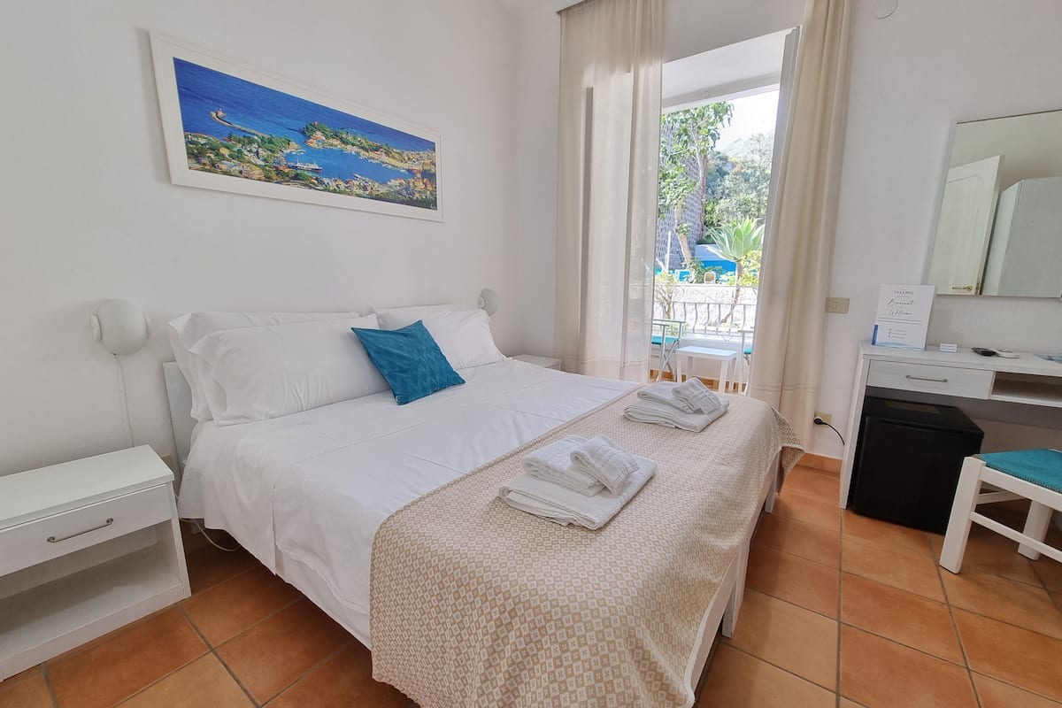 Ein ordentlich eingerichtetes Hotelzimmer mit einem Bett, einem Schreibtisch und einem Stuhl. An der Wand hängt ein Gemälde, ein Fenster mit Vorhängen bietet Blick ins Freie.