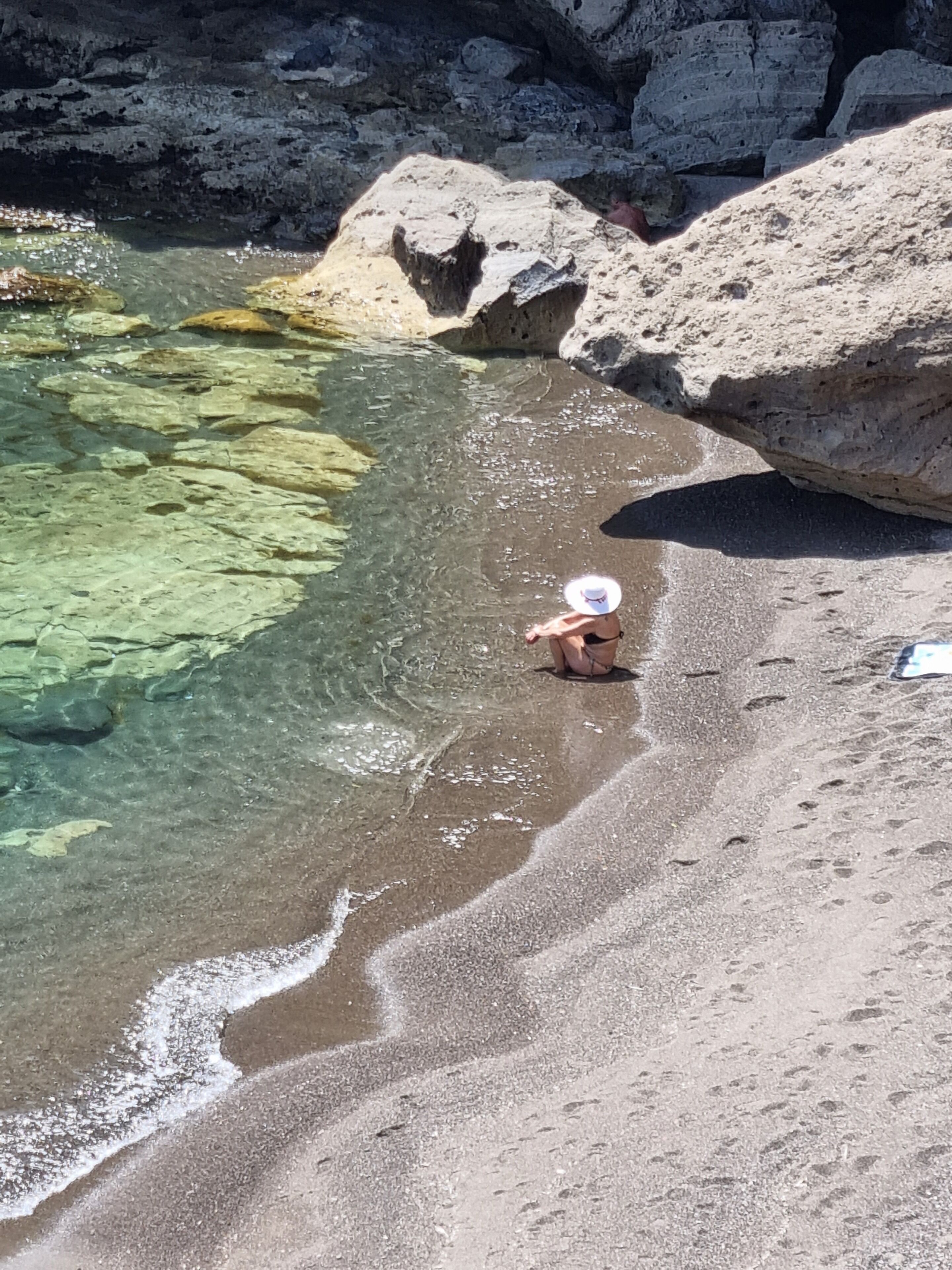 Una playa con aguas claras y poco profundas, formaciones rocosas y una persona con sombrero blanco.