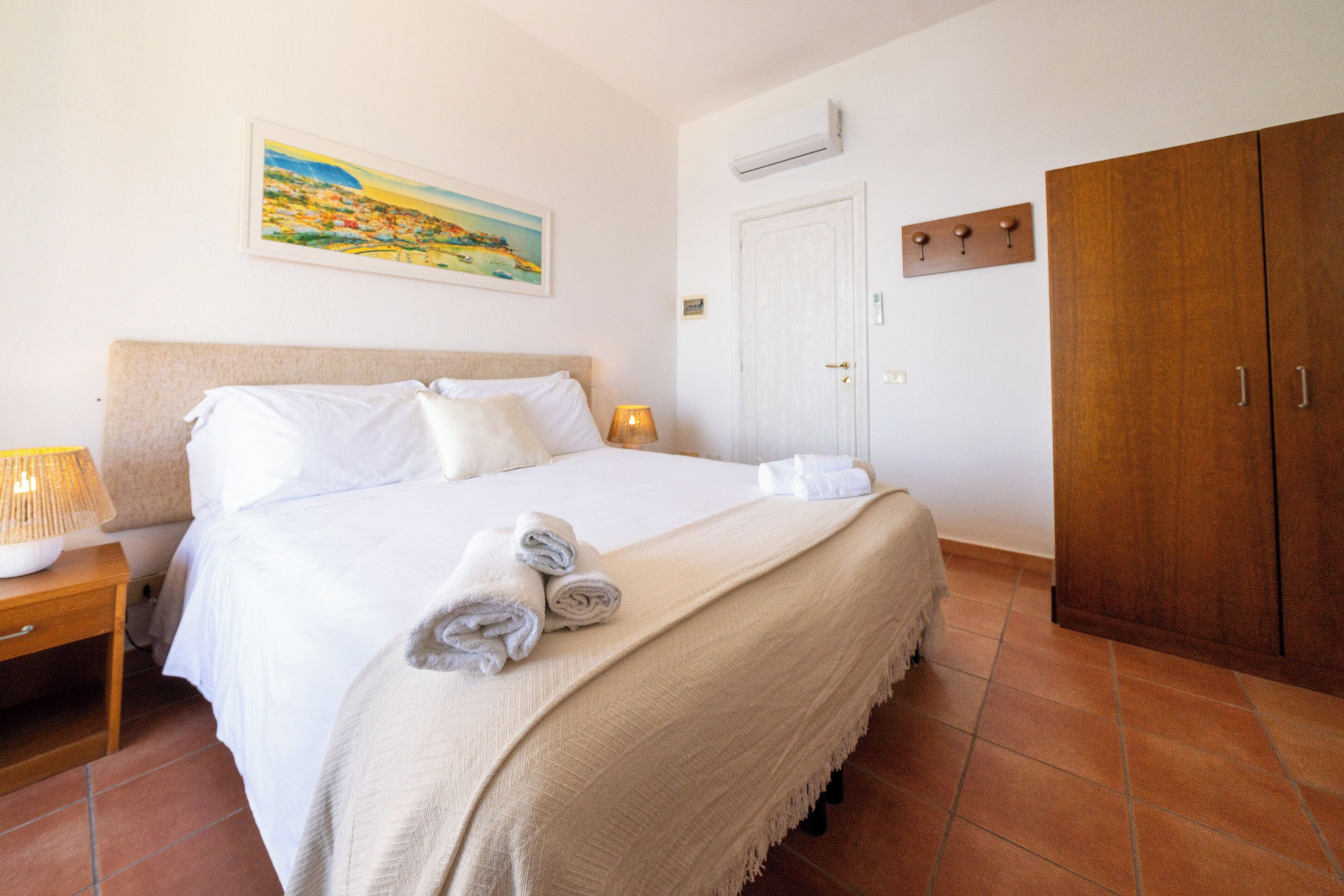 Quarto de hotel com cama, mesa de cabeceira, luminária, um quadro na parede, guarda-roupa e ar condicionado.