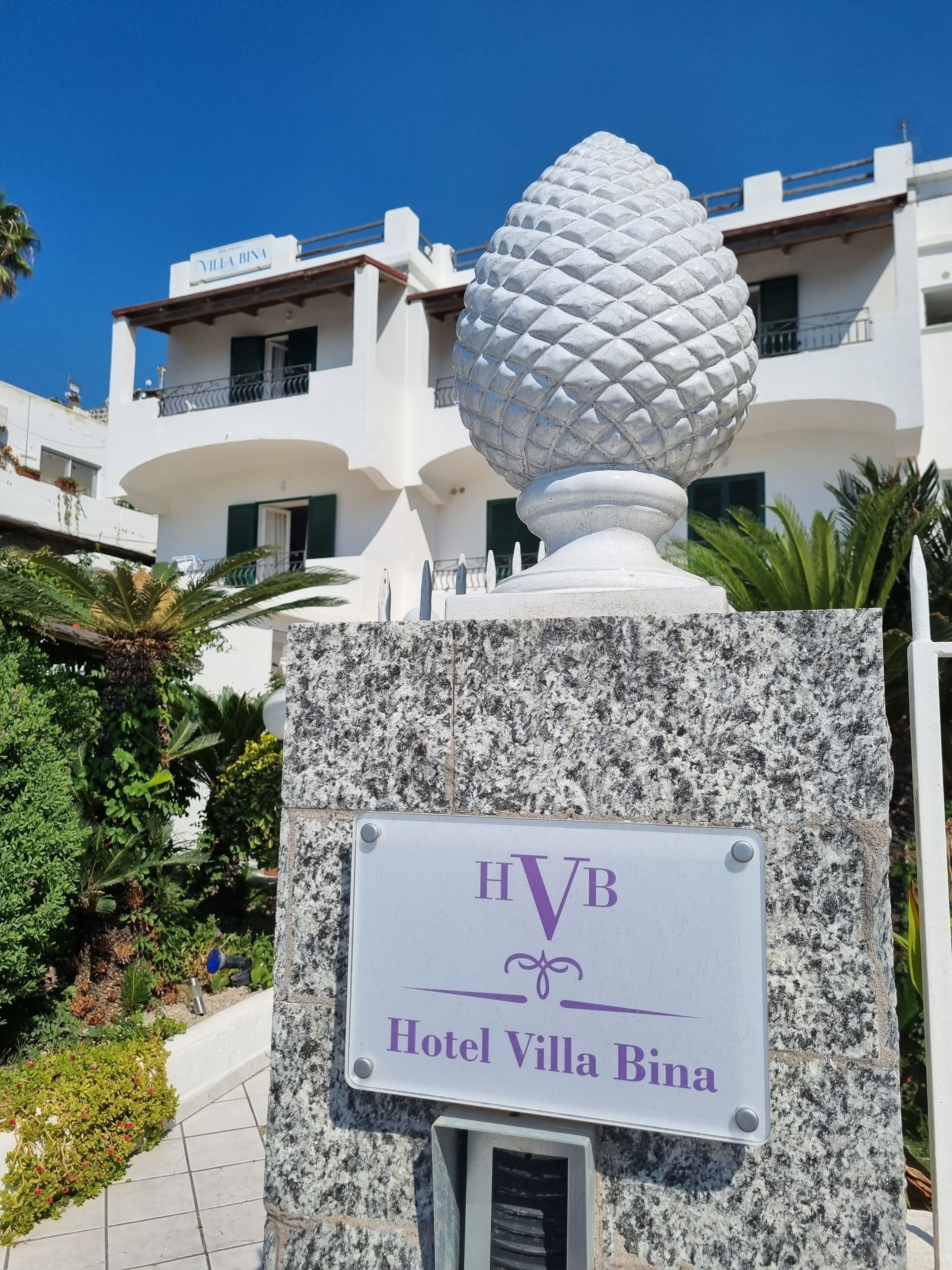 A sign for Hotel Villa Bina with a decorative stone sphere and a clear view of the building in the background.