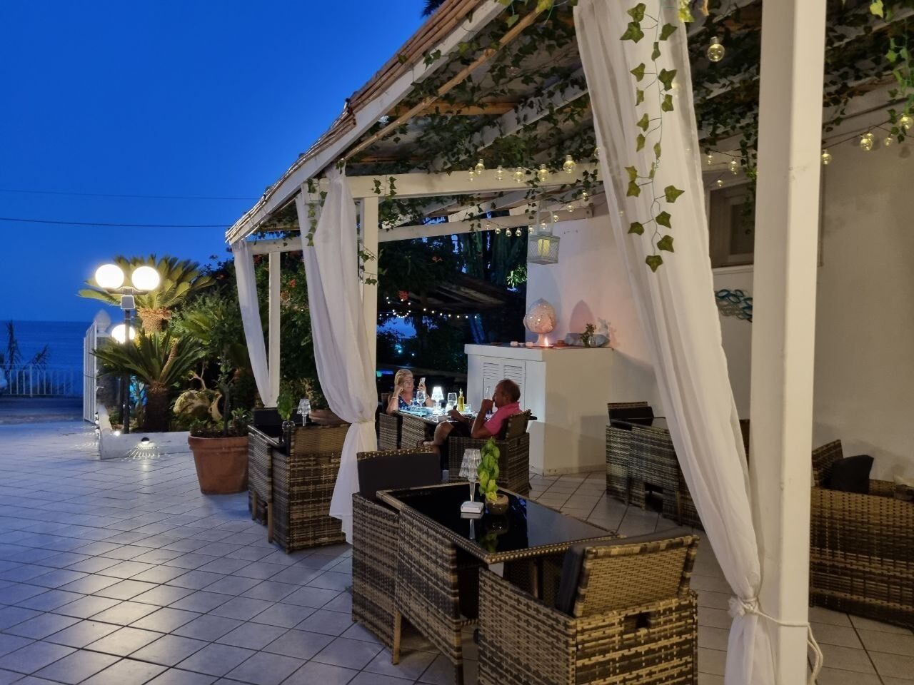 An outdoor patio area with wicker furniture and white drapes, featuring a view of the sea and a bar area.