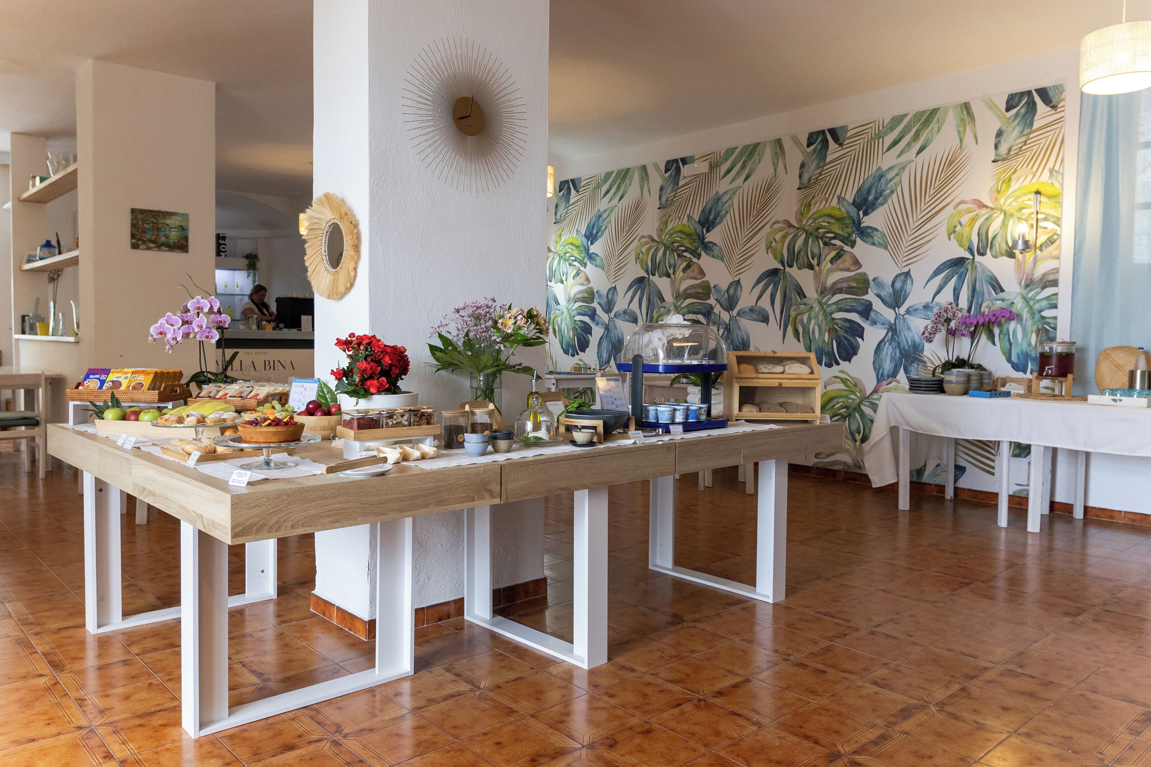 A dining area with a long table set with various dishes, a tropical leaf-patterned wall, and a clock on the wall.