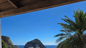 Un balcon avec des fauteuils en osier et une table, offrant une vue sur la côte, avec un palmier et une île rocheuse.