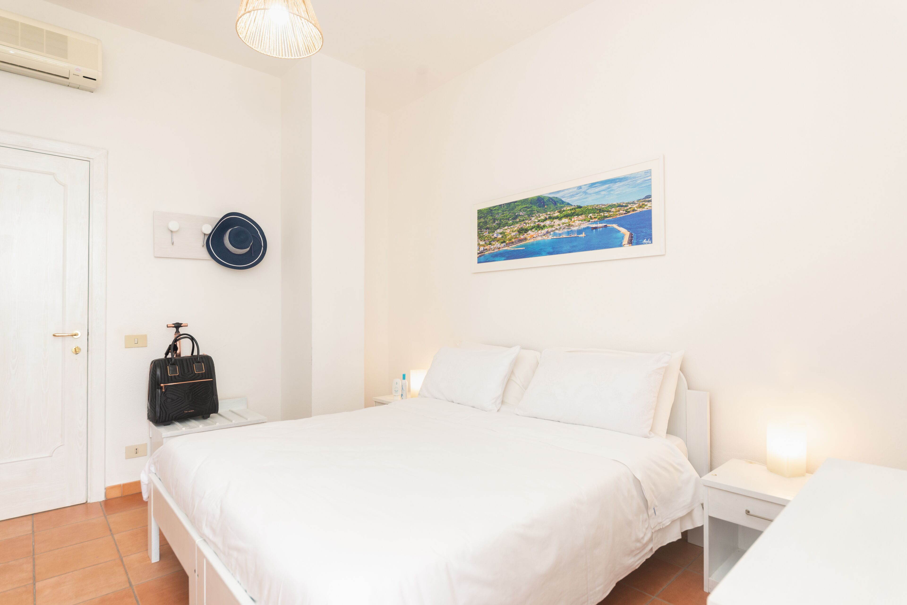 A bedroom with a bed, bedside tables, a nightstand, a wall-mounted air conditioner, a wall-mounted light fixture, a framed picture, and a black handbag on the nightstand.