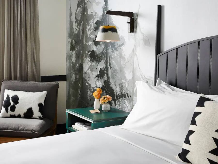 Habitación de hotel con cama, una silla, mesita de noche con lámpara y vistas al exterior.
