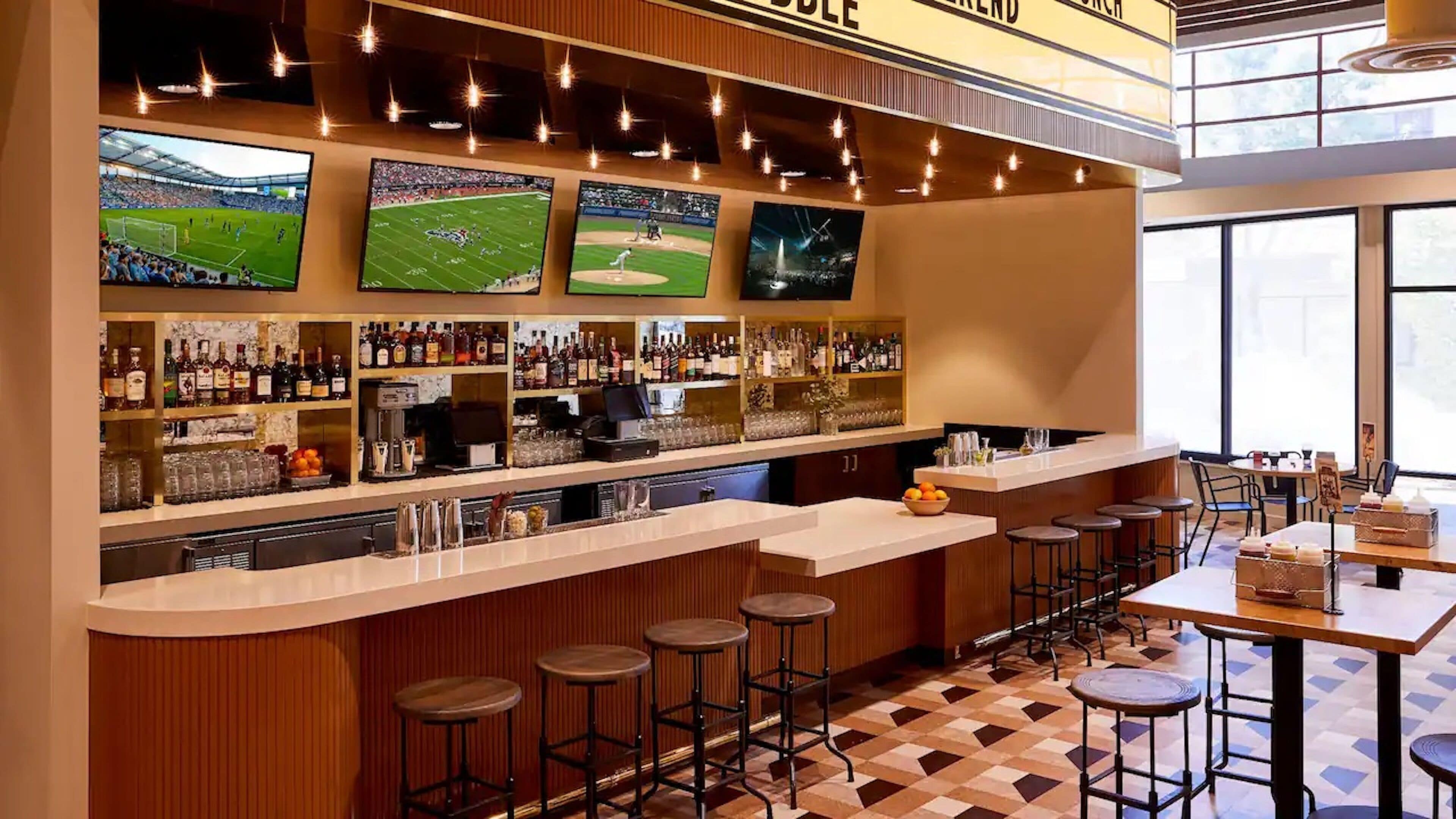 A bar area with multiple screens displaying sports events, a variety of liquor bottles, and stools.