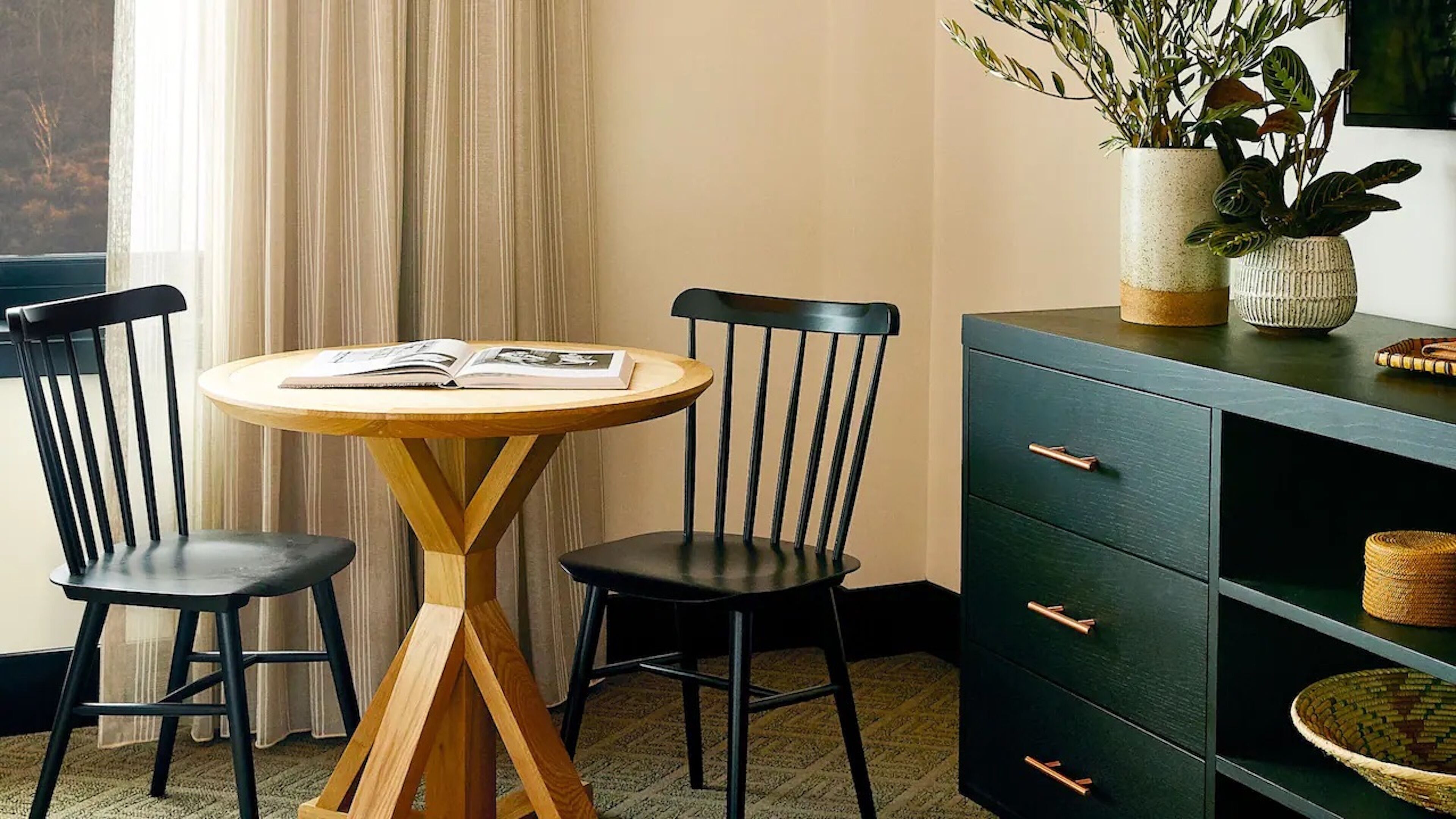 A room with a round wooden table, two black chairs, a dark green cabinet with drawers, and a window with curtains.
