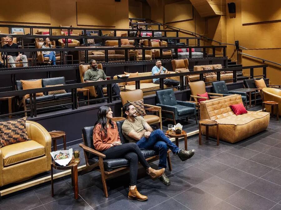 A spacious indoor seating area with tiered rows of chairs and couches, a staircase, and a stage.