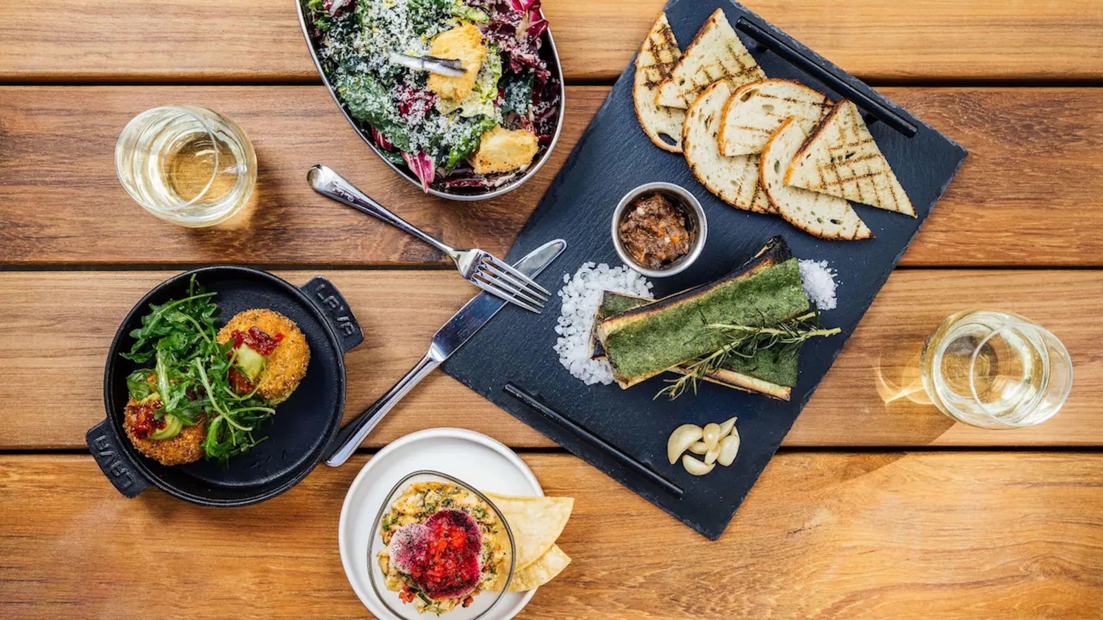 Ein Holztisch mit verschiedenen Speisen, darunter ein Salat, gegrilltes Brot und eine Suppenschale.