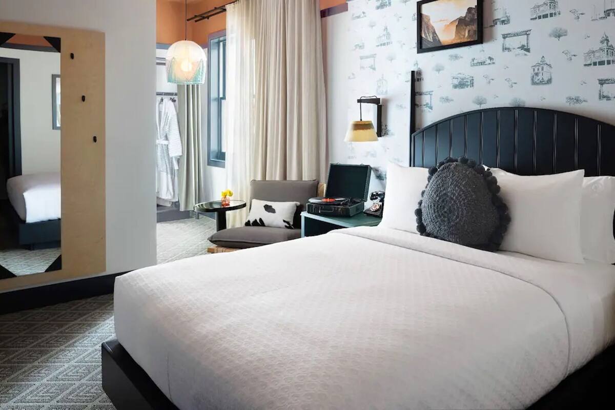 A hotel room with a large bed, a bedside table with a lamp, a mirror, and a wall with a cityscape wallpaper.