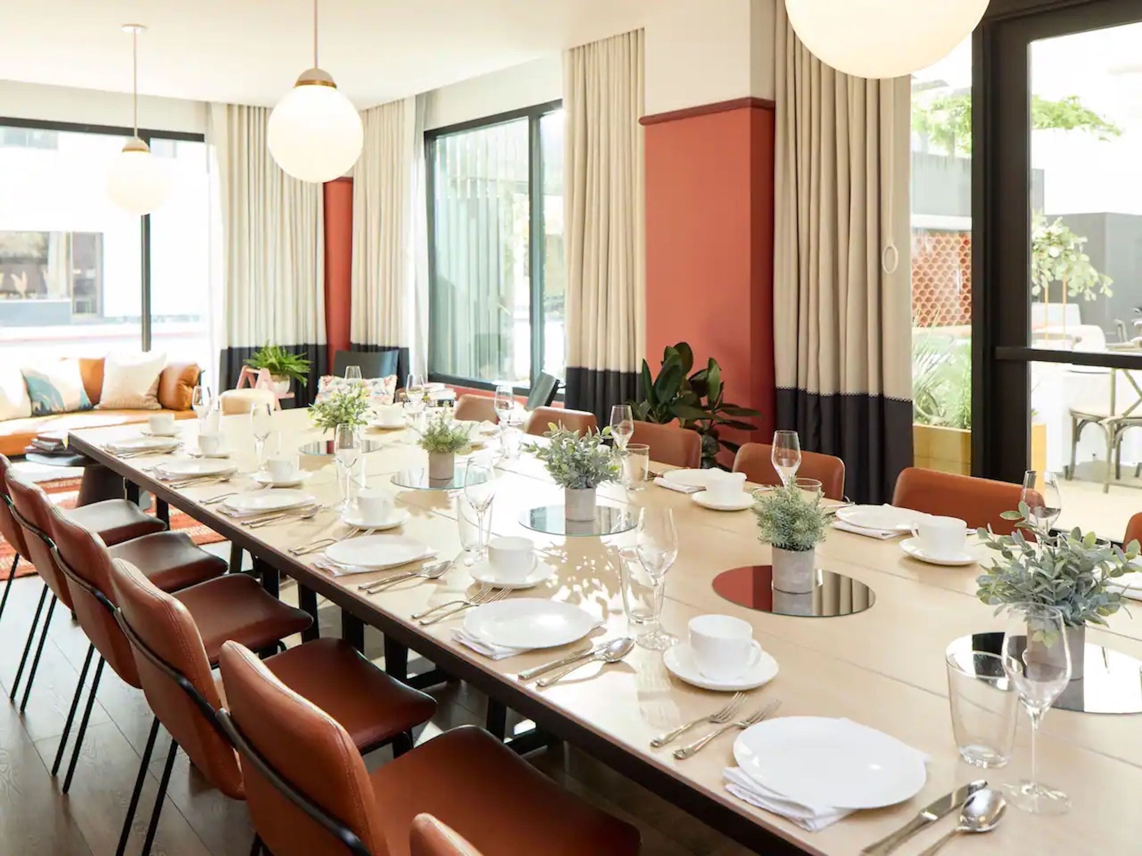 A long dining table set for a meal with multiple chairs, large windows, and curtains.
