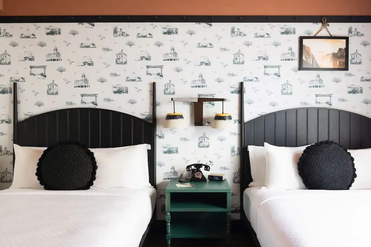 A hotel room with two beds, each with a black headboard and white bedding. There is a green nightstand between the beds with a telephone and a small table. The walls have a pattern of black and white illustrations.