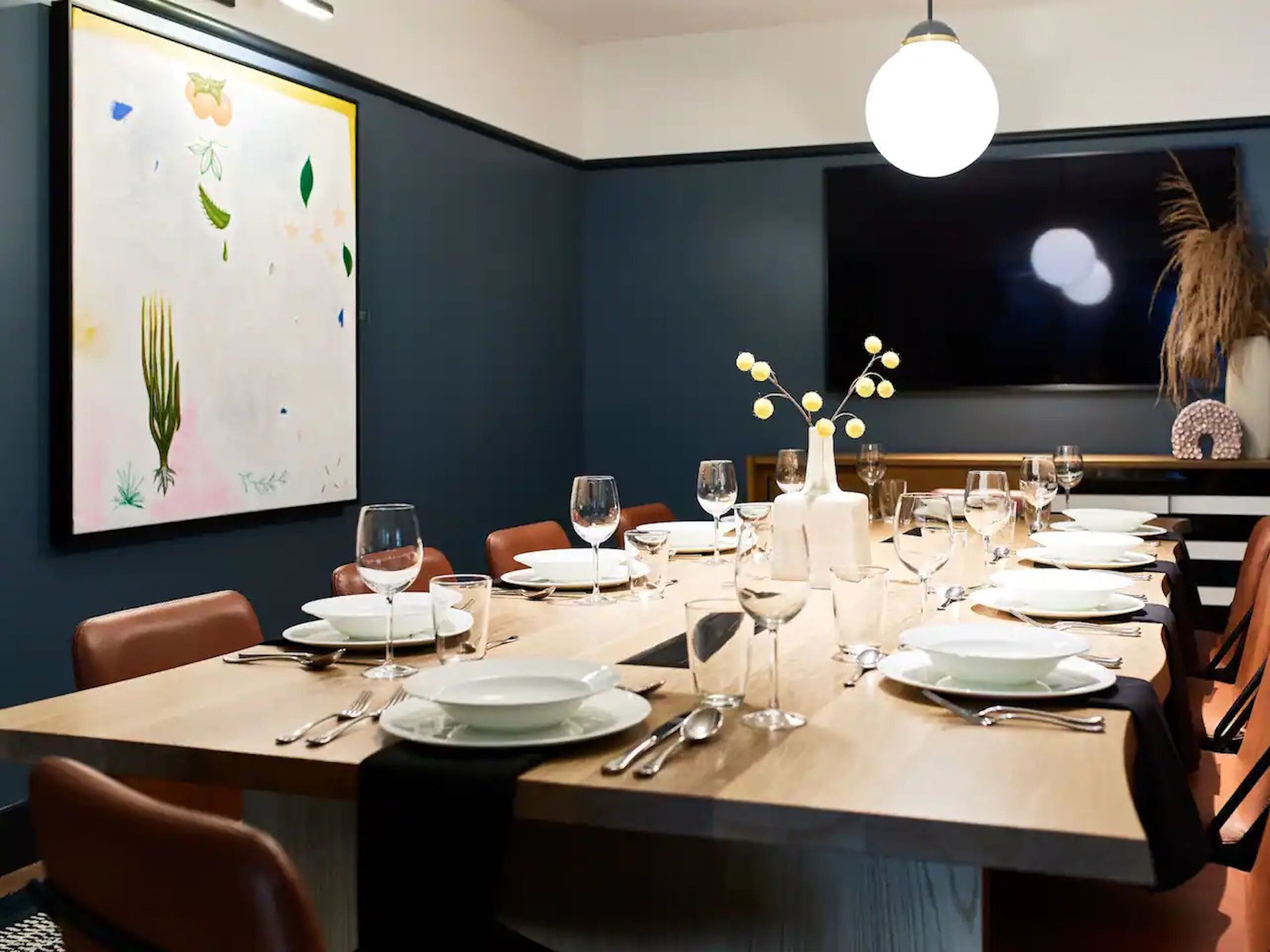 Een eetkamer met een lange tafel gedekt voor een maaltijd, een groot abstract schilderij aan de muur en een televisie aan de andere muur.