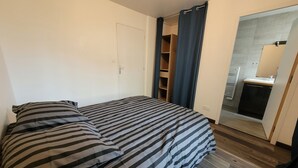 Une chambre à coucher avec un lit, une armoire, une salle de bain visible à travers une porte ouverte et un plancher en bois.