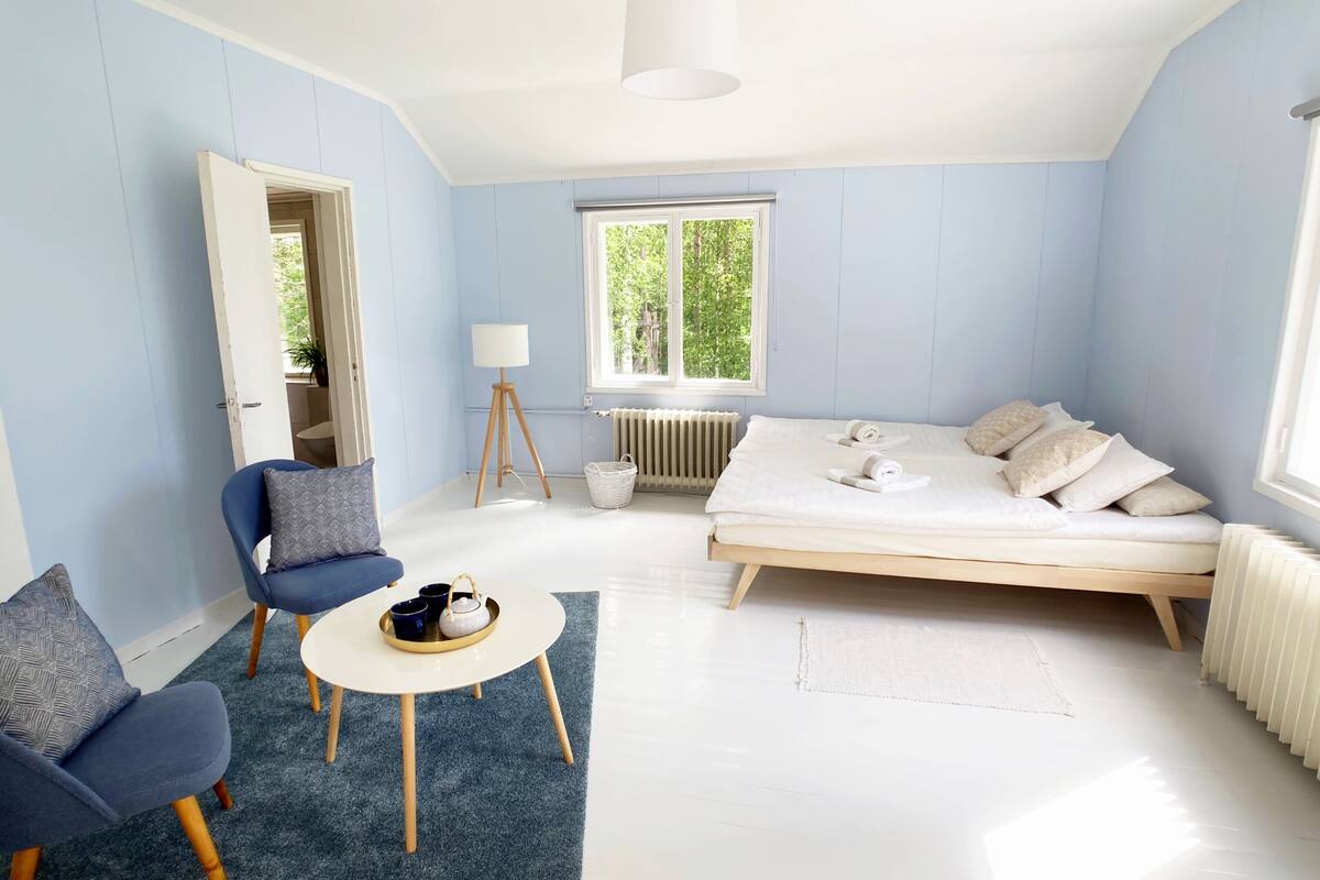 A modern, well-lit room with a bed, a chair, a small table with a tray, and a window providing natural light.