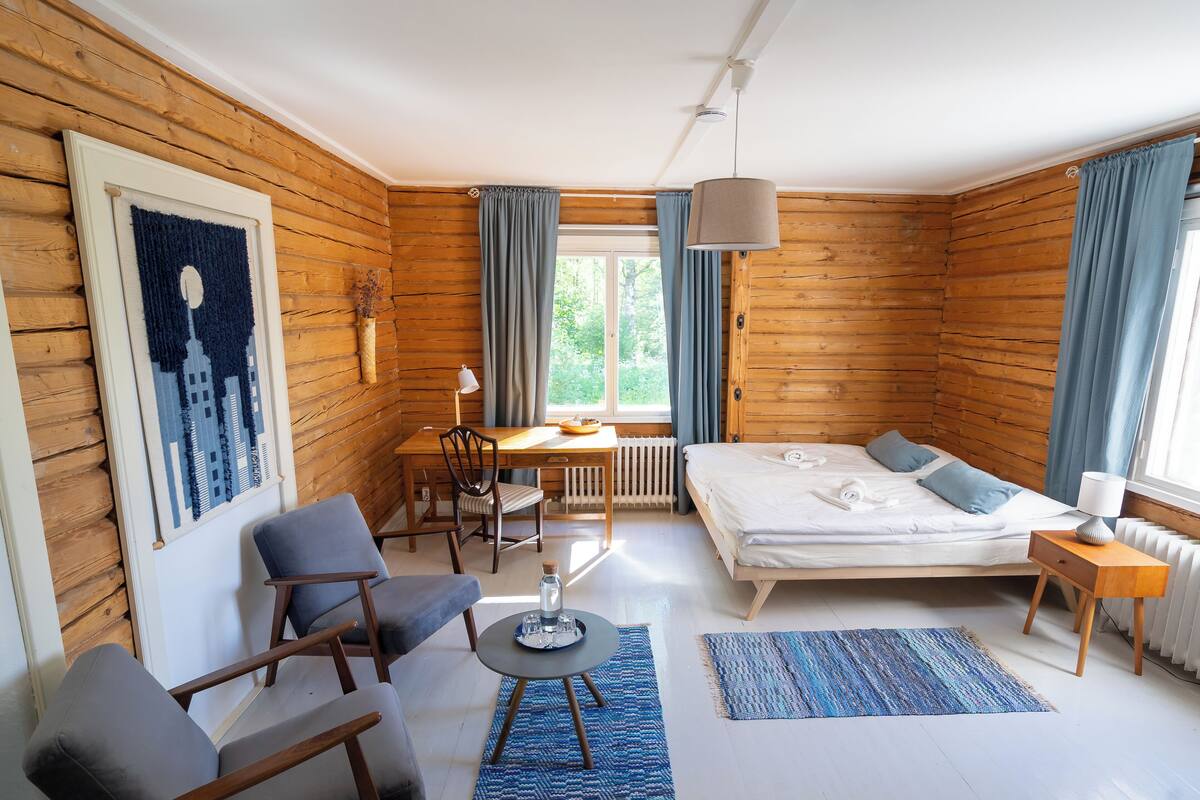 A room with wooden walls, a bed, a desk, a chair, a lamp, and a window with curtains.