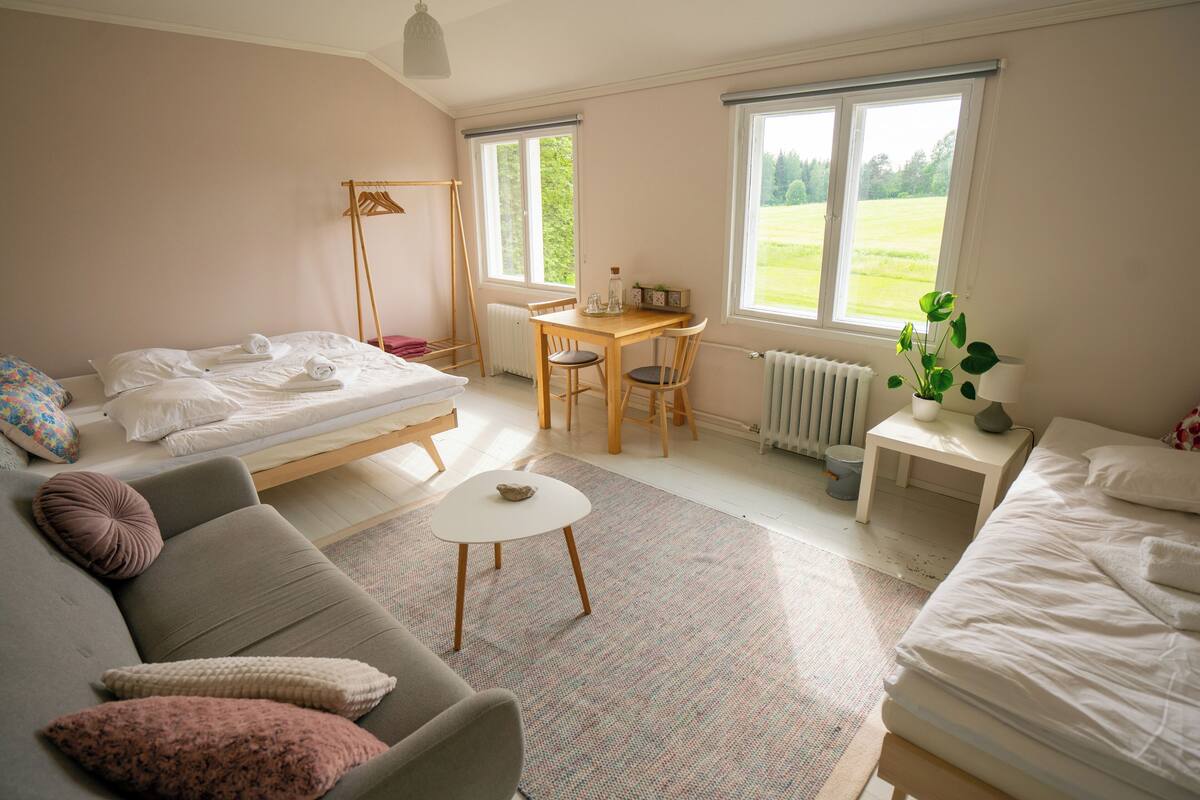 A room with a bed, a sofa, a small table, and a chair. There are windows with a view of a green field.