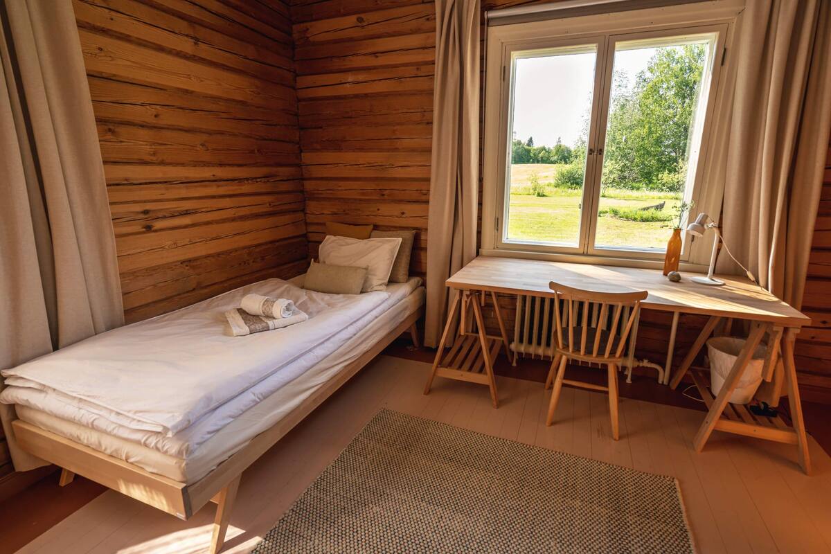 A wooden cabin room with a bed, a table, and a window overlooking a garden.