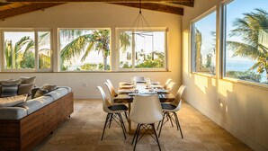 A dining area with a wooden ceiling, large windows with palm trees, a dining table set with chairs, and a sofa with cushions. - Private Villas with Amazing Views - Private Pools, 6 King Bedrooms @ Villa Topaz (West Bay)