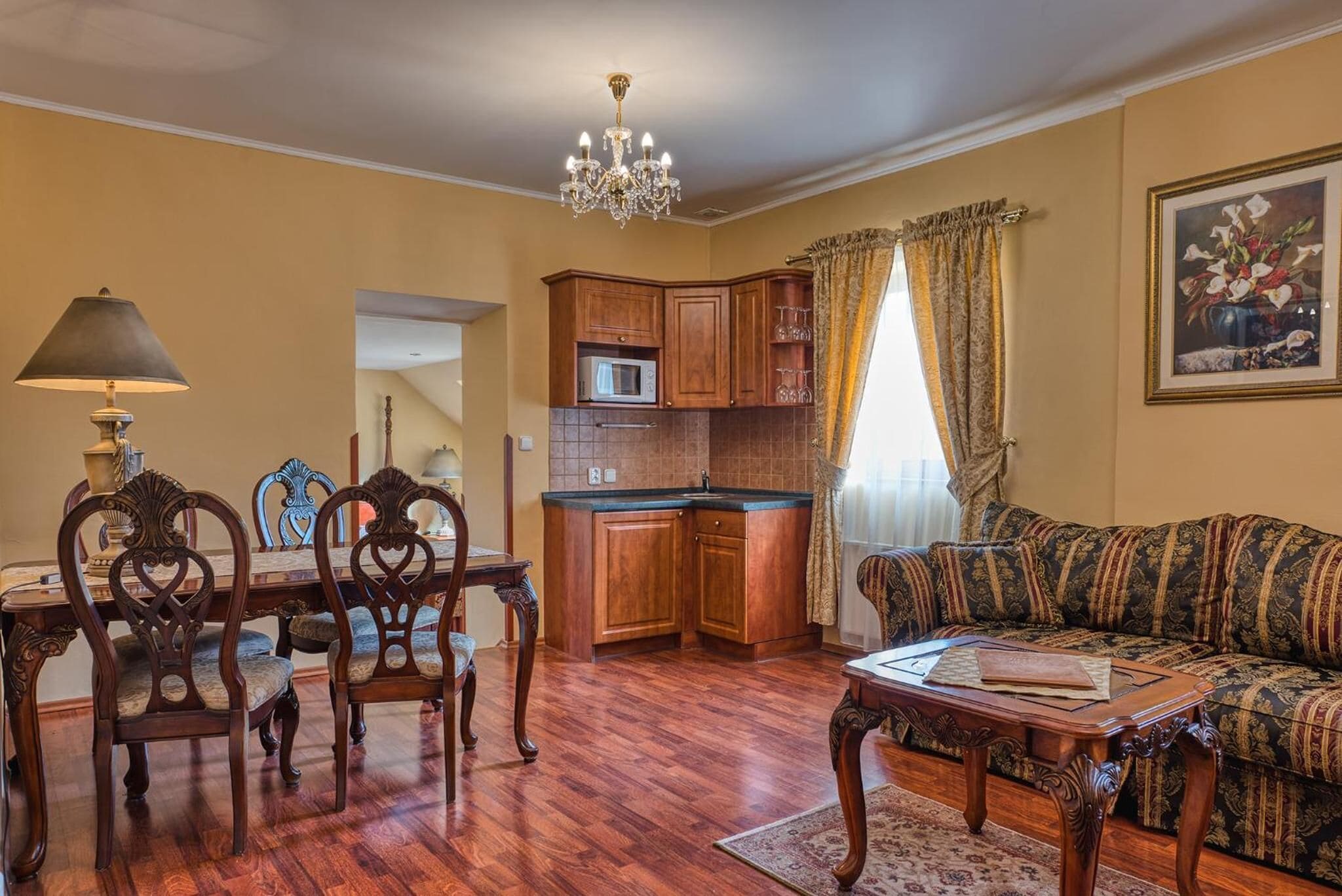 A living room with a dining table, chairs, a sofa, a chandelier, and a kitchen area with cabinets and a microwave.