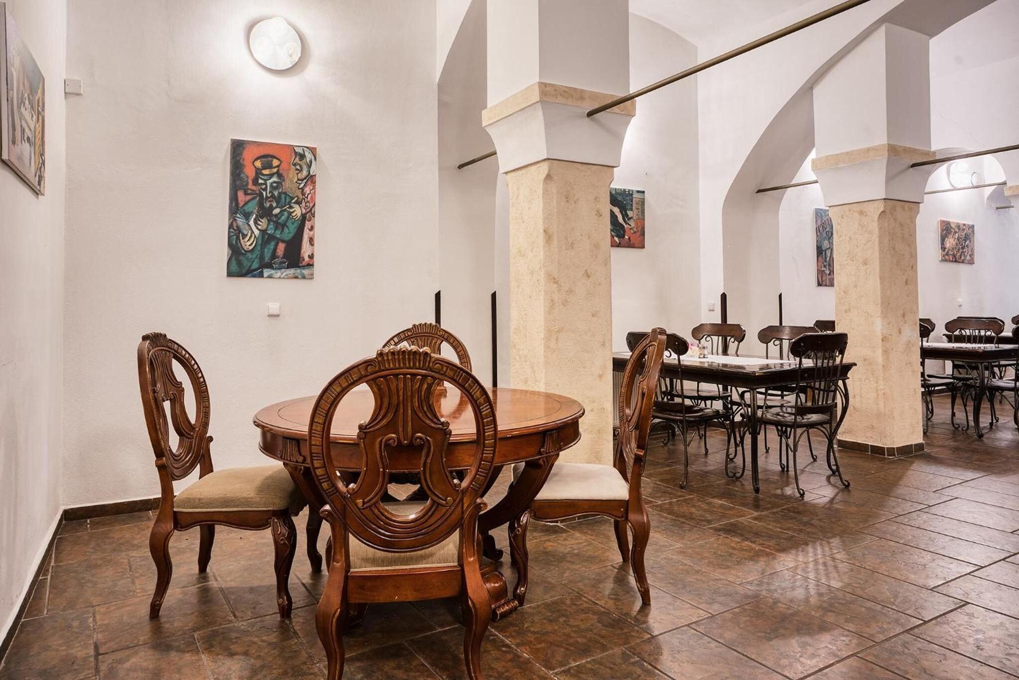 Comedor con sillas de madera decoradas y una mesa redonda, suelo de baldosas y paredes blancas con obras de arte.