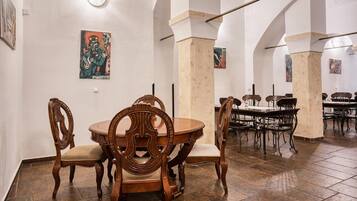 Comedor con sillas de madera decoradas y una mesa redonda, suelo de baldosas y paredes blancas con obras de arte.