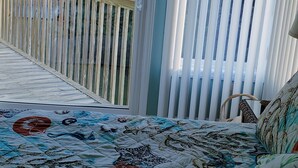 A balcony with a wooden railing, a bed with a quilted bedspread, and a view of the ocean and buildings. - "A Lil' Nauti" - 2 Bedroom Condo - Ocean Views, Emerald Isle, NC (Emerald Isle)