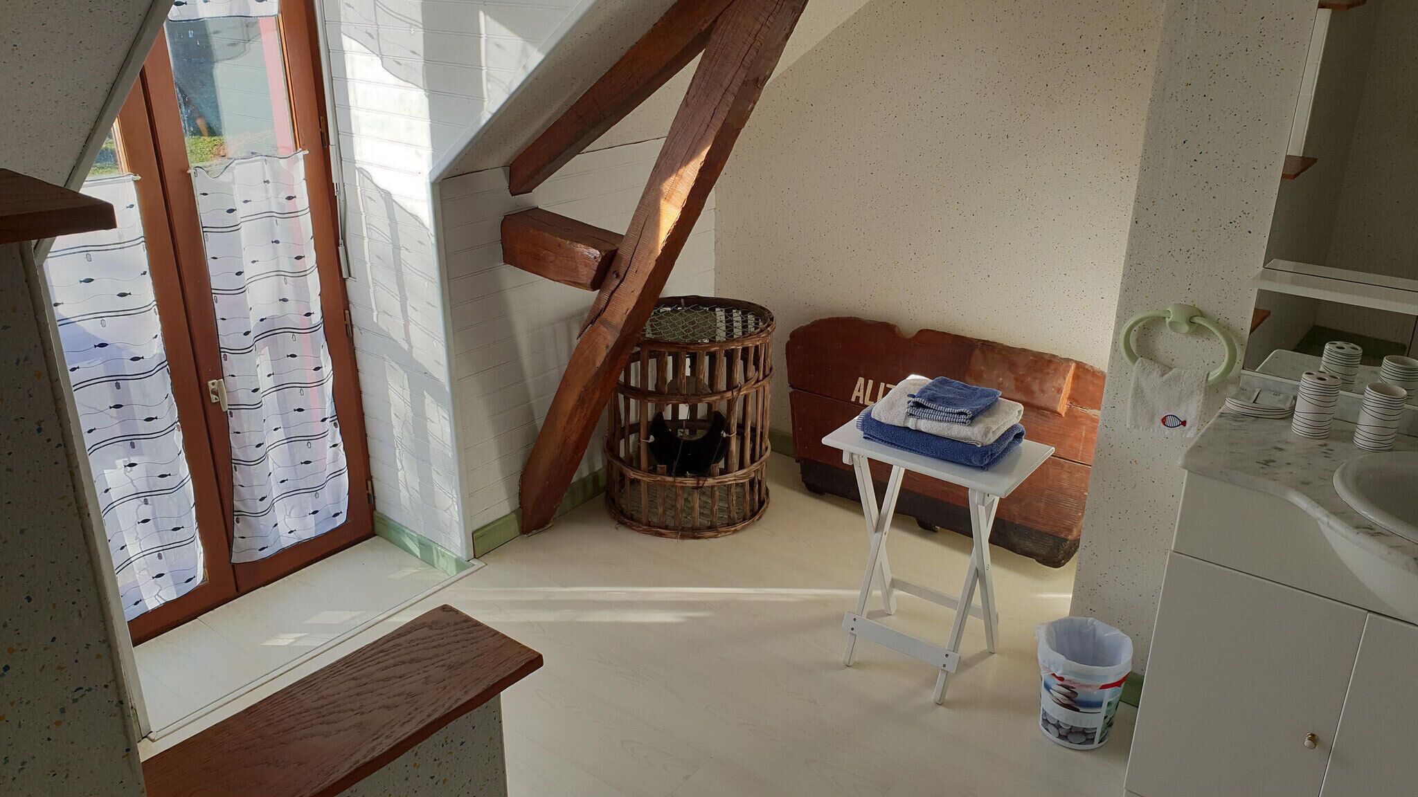 A small, compact kitchen with a window, a wooden stool, a small table, and a trash bin.