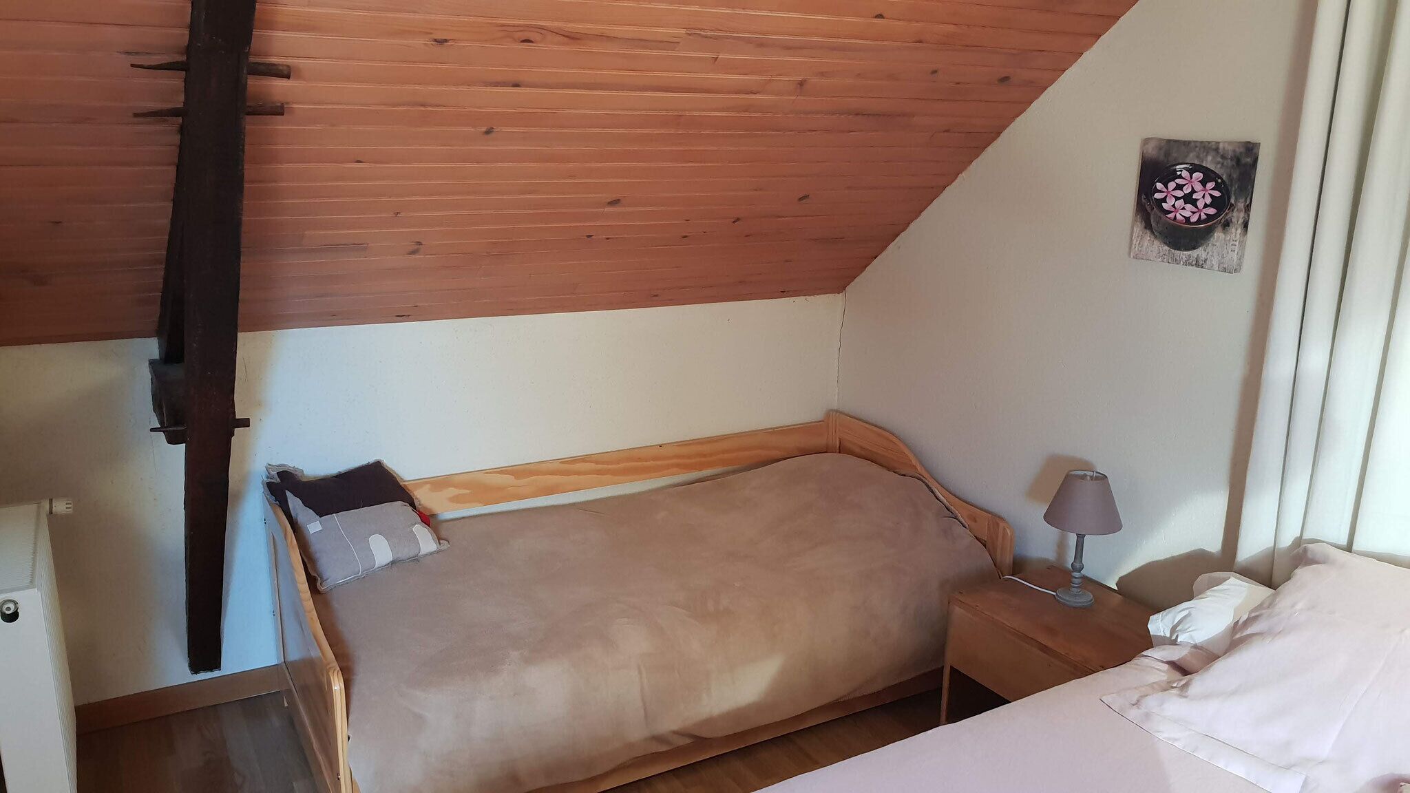 A small, cozy room with a wooden ceiling, a single bed with a wooden headboard, a nightstand with a lamp, and a wall-mounted picture.