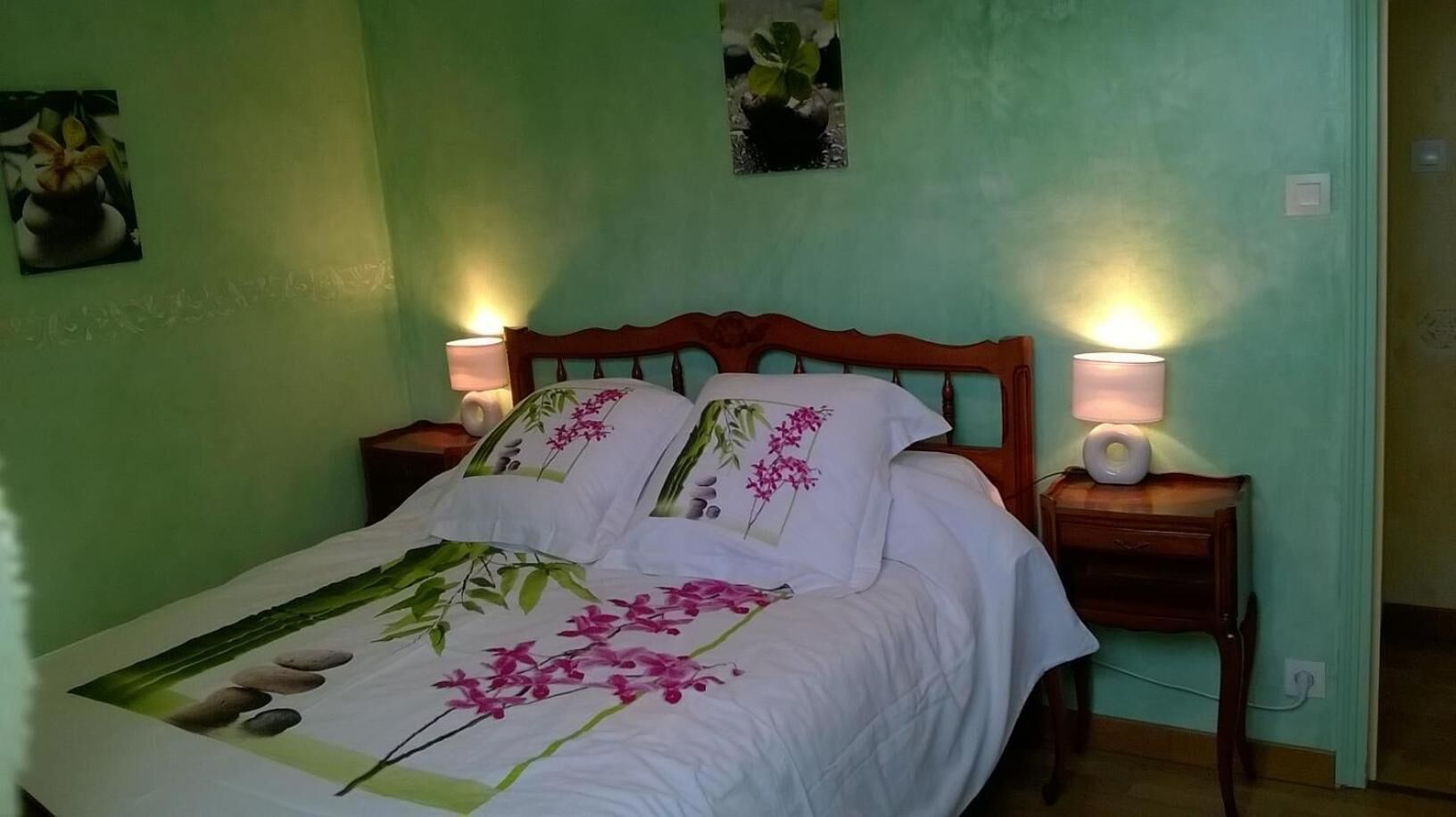 Ein ordentlich bezogenes Bett mit floralem Bettbezug, zwei Nachttischlampen und ein kleiner Nachttisch mit Lampe.