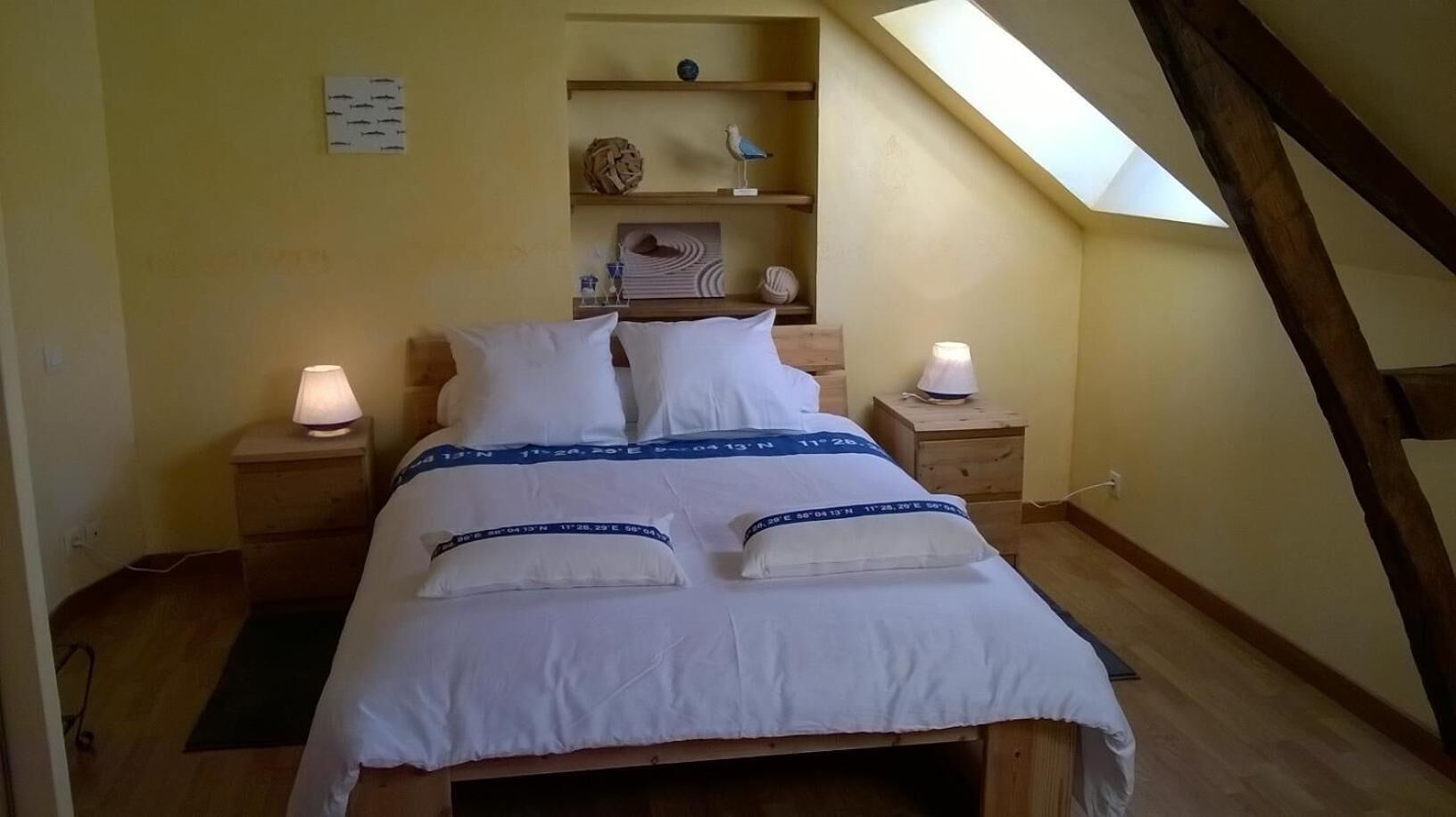 A bedroom with a skylight, wooden beams, a bed with white bedding and blue accents, two bedside tables with lamps, and a shelf with decorative items.