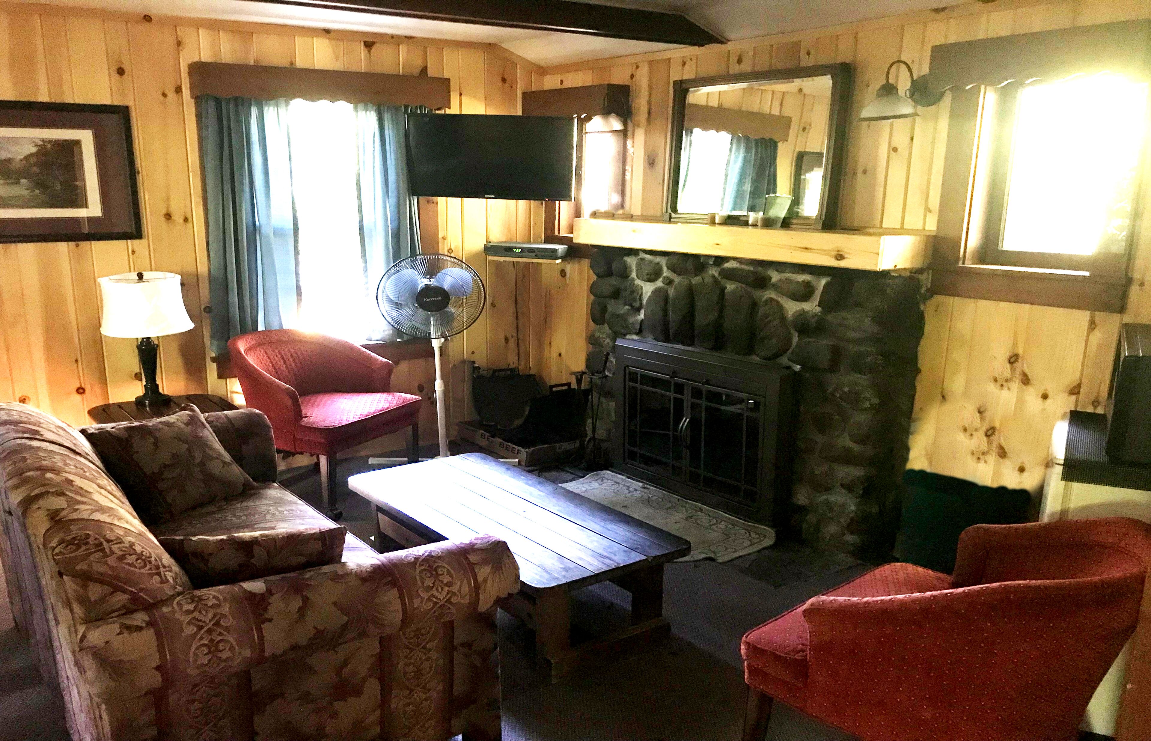 A cozy living room with a stone fireplace, wooden walls, a television, and a fan.