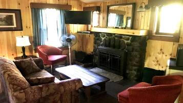 A cozy living room with a stone fireplace, wooden walls, a television, and a fan.