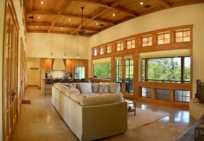 Amplia sala de estar con un sofá modular grande, vigas de techo de madera y grandes ventanales que ofrecen vistas a zonas verdes.